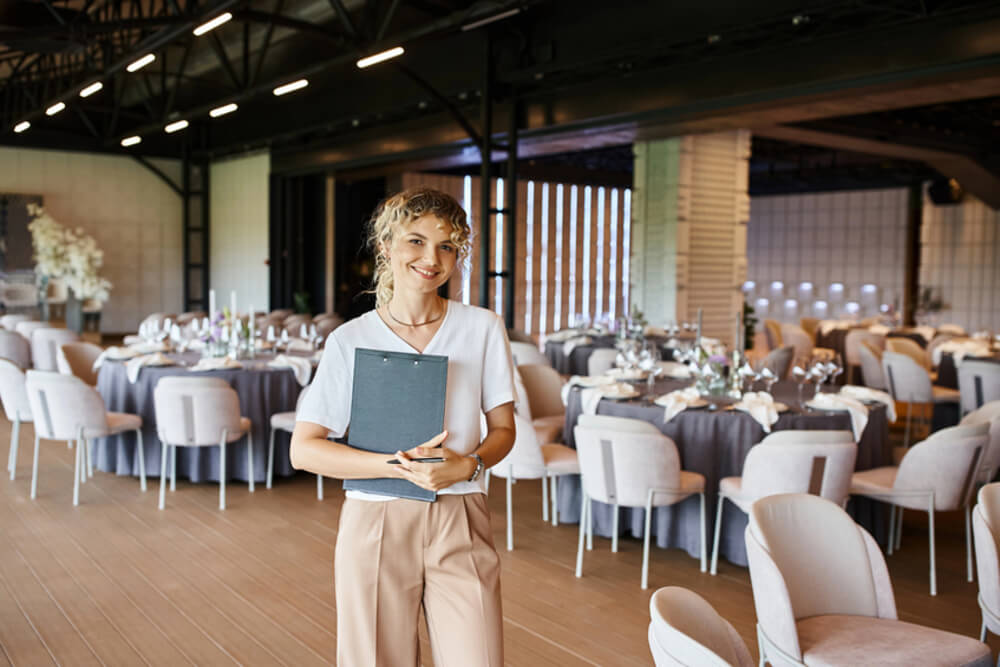 Woman with curly blonde hair in a low ponytail stands smiling with a clipboard in a room full of round banquet tables set with flowers and plates.