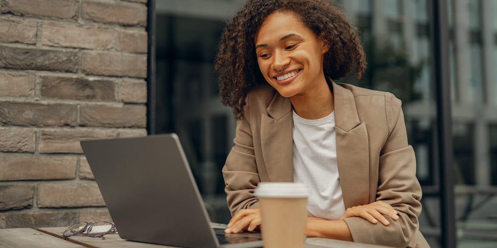 A person sitting at a table with a cup of coffee, smiling while using a laptop.