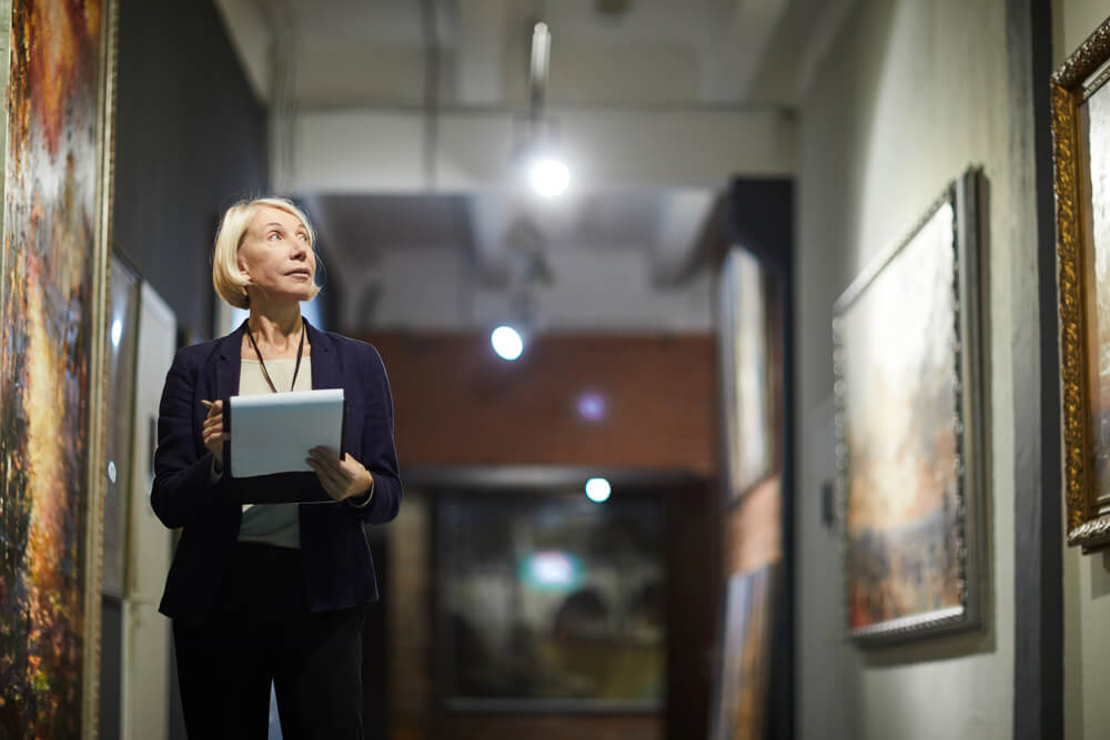 A blonde woman stands in a hallway of art looking up at an artwork with a notebook in her hands.
