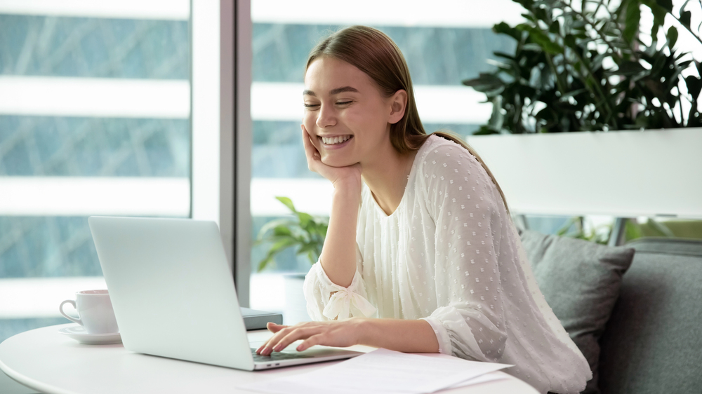 Woman sits smiling while looking at her laptop. Her face is in her right hand.